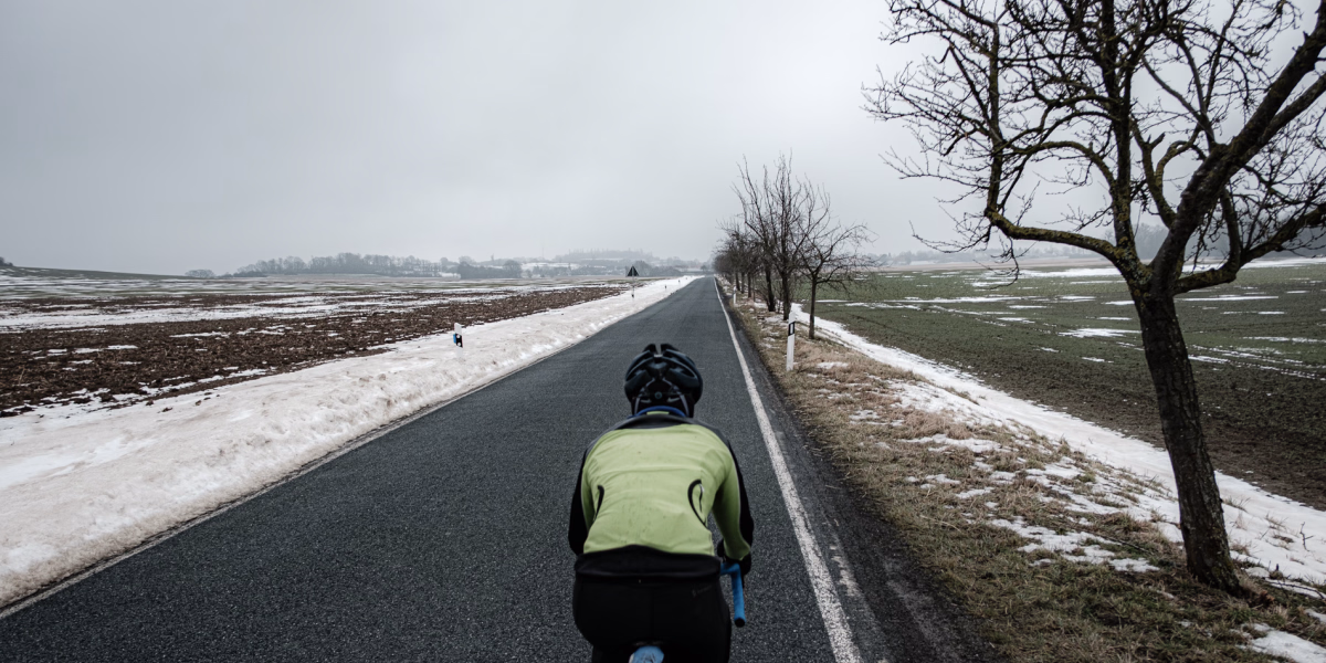 Radsportlerin von hinten zu sehen fährt auf einer schnurgraden Landstrasse Richtung Stemmen. Schnee, rechts und links der Strasse, Obstbäume auf der rechten Seite. Der Stemmer Berg im Hintergrund.