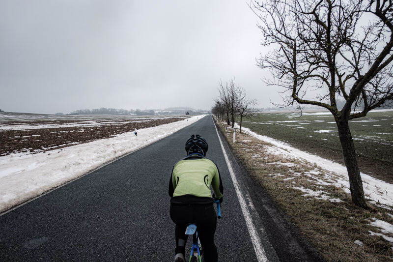Radsportlerin von hinten zu sehen fährt auf einer schnurgraden Landstrasse Richtung Stemmen. Schnee, rechts und links der Strasse, Obstbäume auf der rechten Seite. Der Stemmer Berg im Hintergrund.
