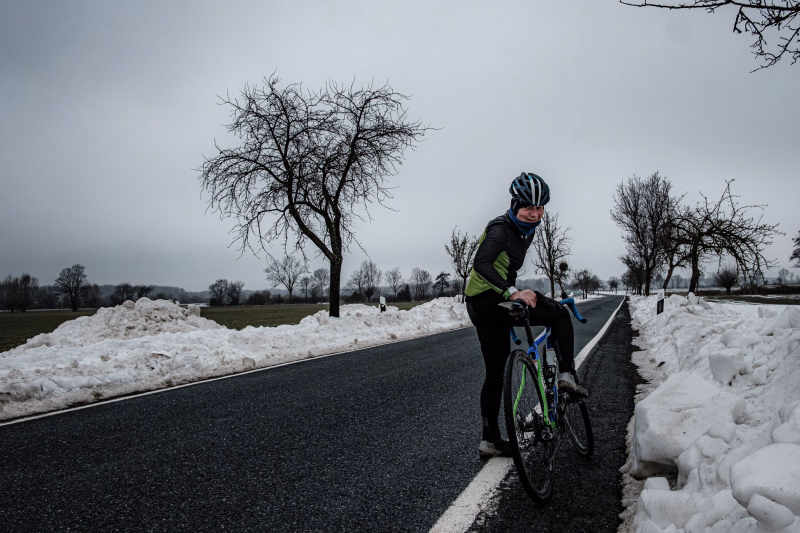 Landstrasse kurz hinter Ditterke, Richtung Kirchwehren, mit viel Schnee an den Seiten und einzelnen Obstbäumen. Eine warm angezogene Radsportlerin steht am Strassenrad und lacht in die Kamera.