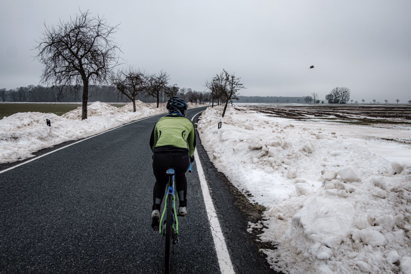 Radsportlerin mit grüner Winterjacke fährt auf der Landstrasse Richtung Kirchwehren und blickt einem Bussard nach, der den Weg gekreuzt hat.. Rechts und links viel Schnee, einzelne Obstbäume am Strassenrand.