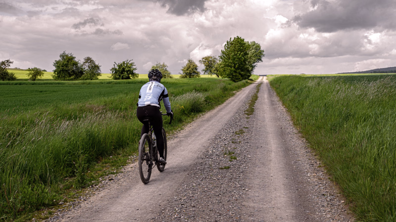Ein grauer, schottriger Feldweg führt gerad auf den goldenen Schnittpunkt hin. Auf dem Feldweg eine Radsportlerin mit Gravelbike von hinten zu sehen. Grüne Getreidefelder rechts und links, im Hintergrund ein paar Bäume.