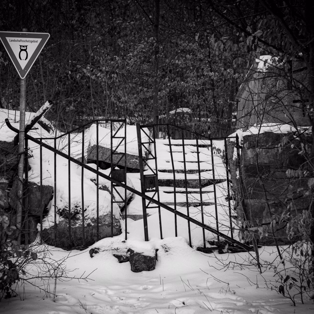Das alte Eingangsportal vom Benther Berg Gasthaus. Verfallene Mauern und Treppen. Schnee auf den Stufen. Das alte Eisentor verrammelt mit einem Eisenrohr. Davor ein Schild Landschaftsschutzgebiet.